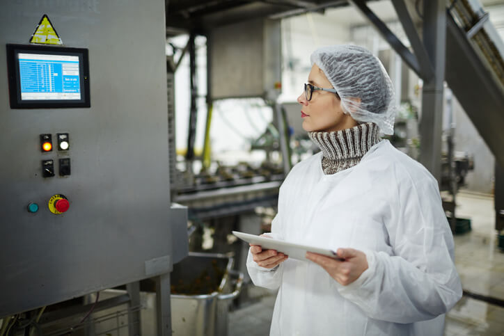 Staff of food factory in protective uniform looking at display with regullation information on wall of industrial machine